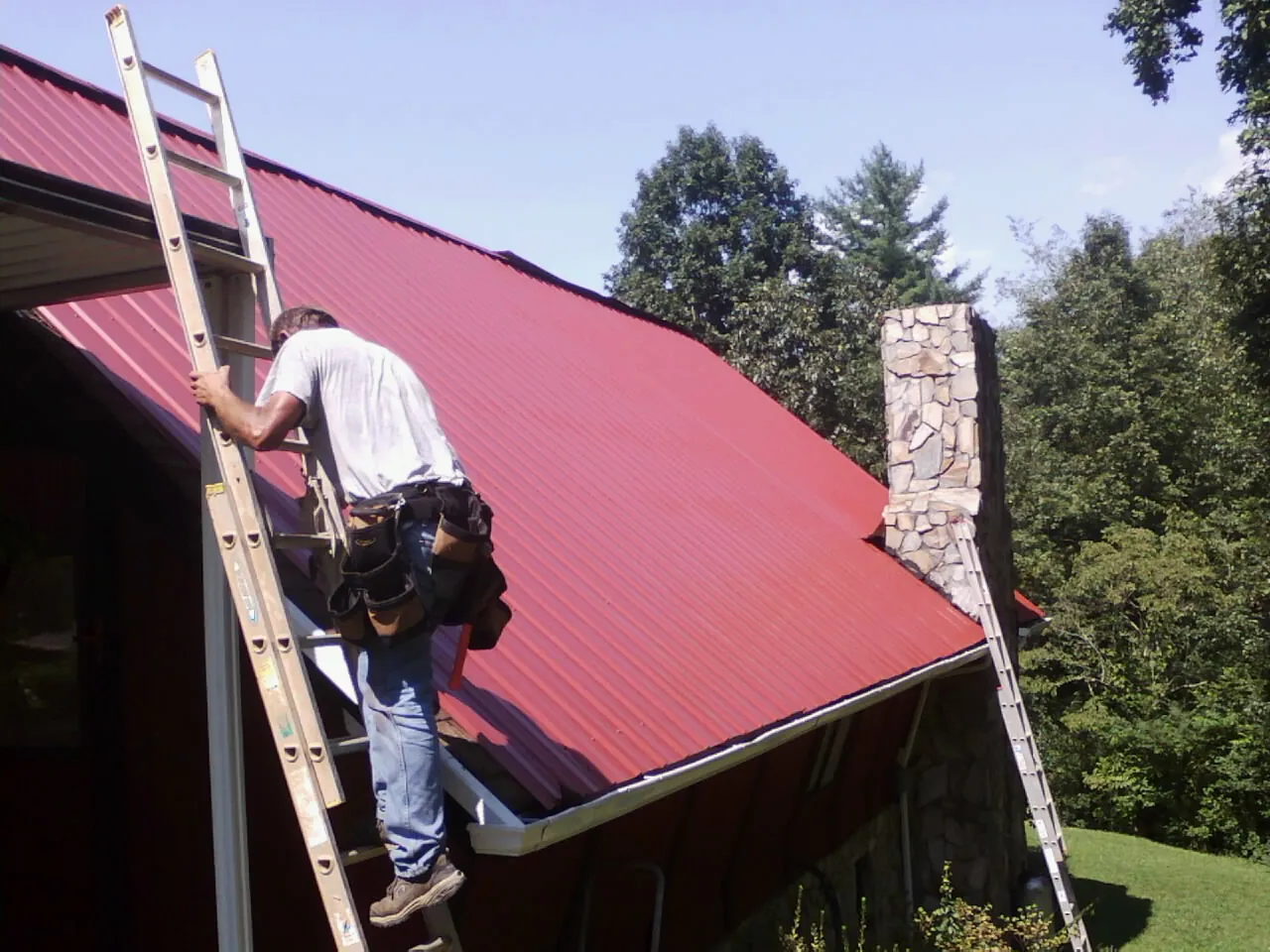 Roofer on a ladder beside a red metal roof and stone chimney — Soffit & Fascia Repair by Roofing Brothers in Murphysboro